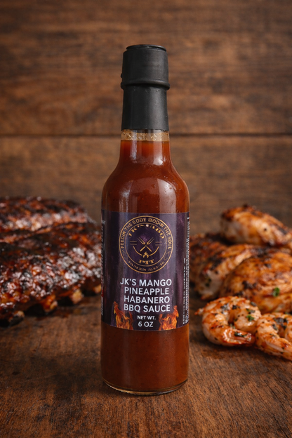 JK’s Mango Pineapple Habanero BBQ Sauce bottle on rustic wooden background with grilled chicken and BBQ ribs
