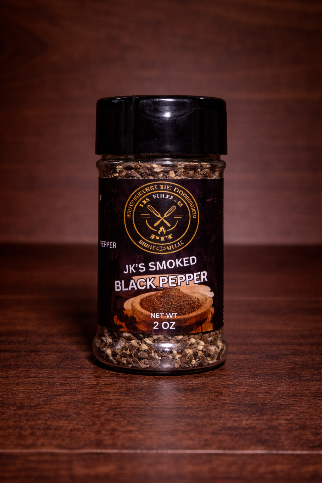 JK’s Smoked Black Pepper 2 oz bottle on wooden background with coarse cracked pepper visible in clear seasoning jar