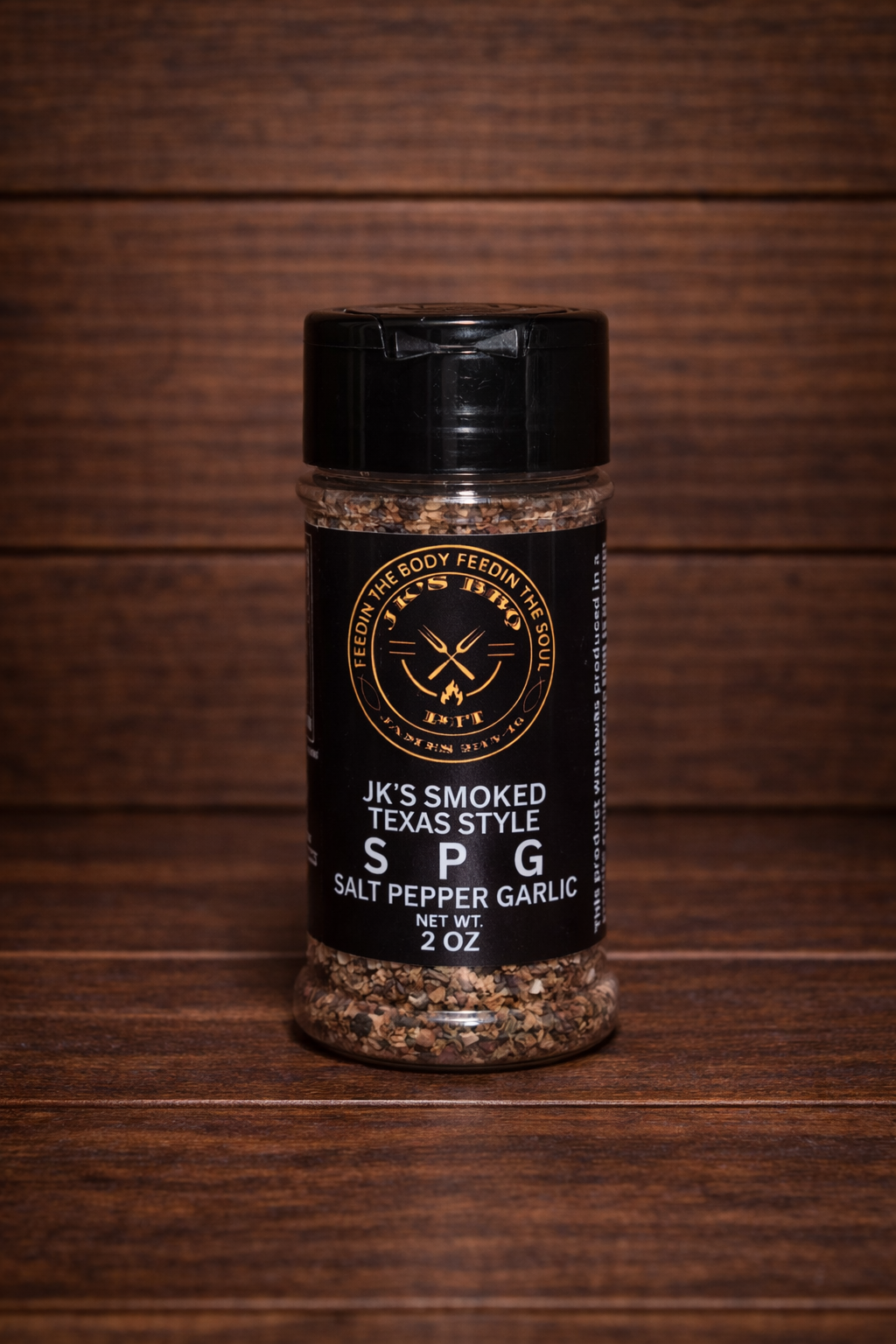 JK's Smoked Texas Style SPG seasoning 2 oz bottle on dark rustic wooden table and wood wall background – salt pepper garlic BBQ rub for brisket, steak, and grilling