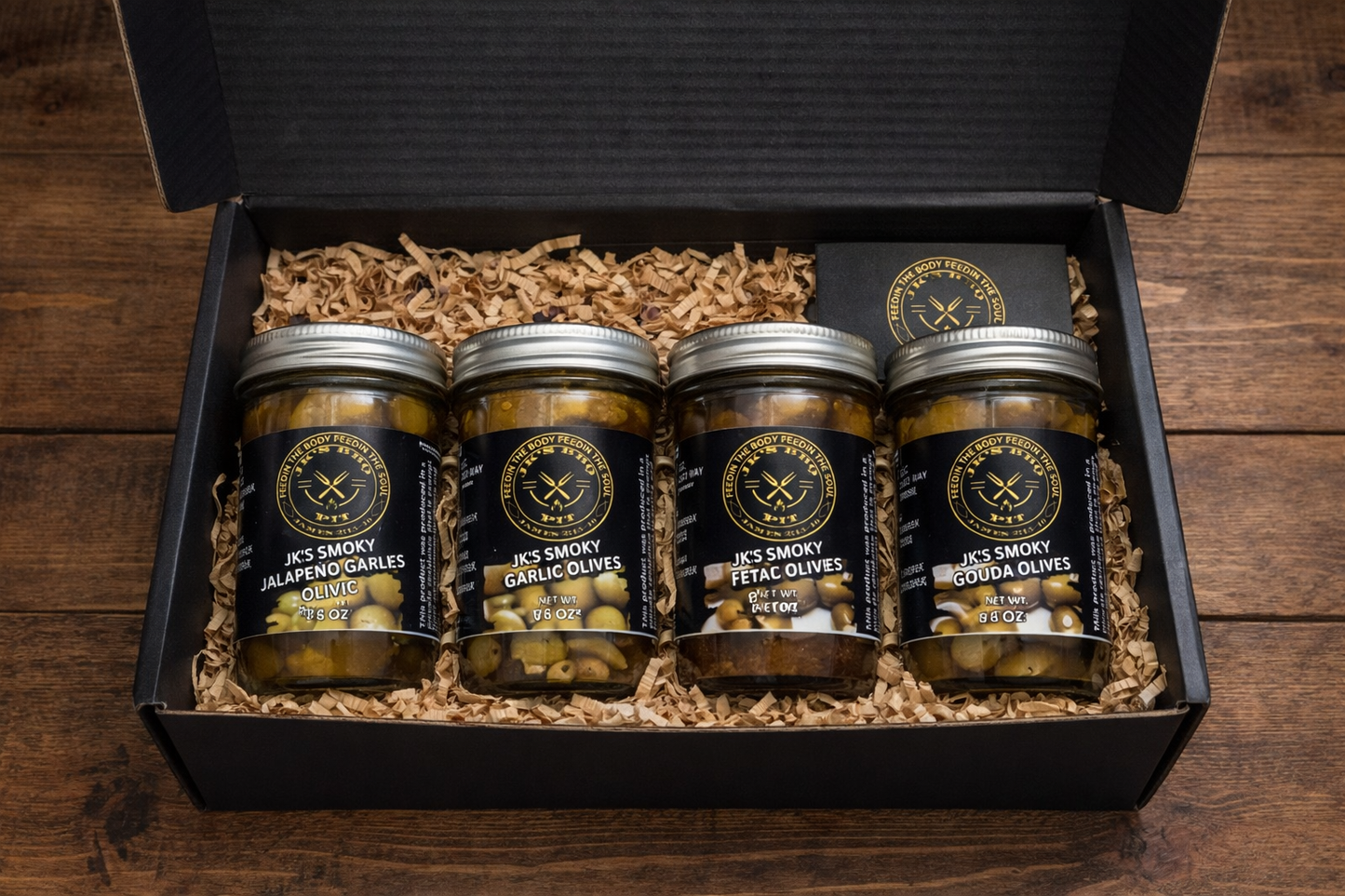 Smoked olive sampler bundle with garlic, gouda, feta and jalapeño olives in black BBQ gift box with wood shavings on wooden background